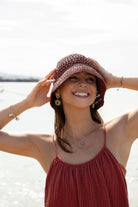 Model smiling and holding the brim of a handwoven raffia hat by the sea