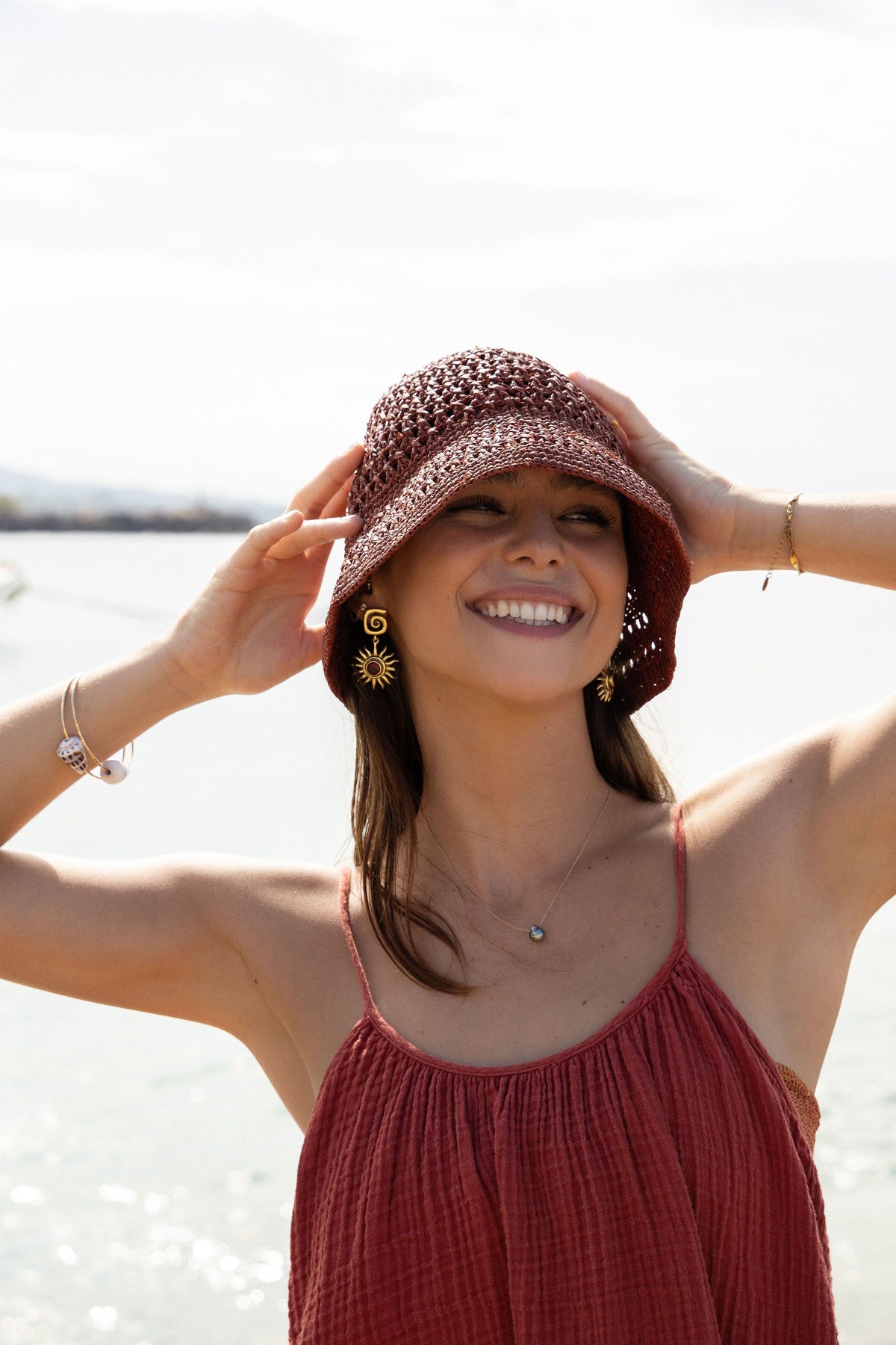 Model smiling and holding the brim of a handwoven raffia hat by the sea